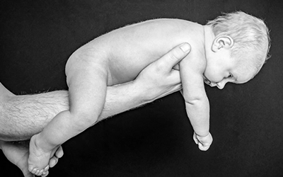 Ein neugeborenes Baby, nackt und zart, liegt längs auf dem ausgestreckten Arm des Vaters. Das Schwarz-Weiß-Foto betont die Geborgenheit und die Größe der Vaterhand im Vergleich zum Säugling. Ein intimer Moment der Babyfotografie, festgehalten von Michael Schalansky.